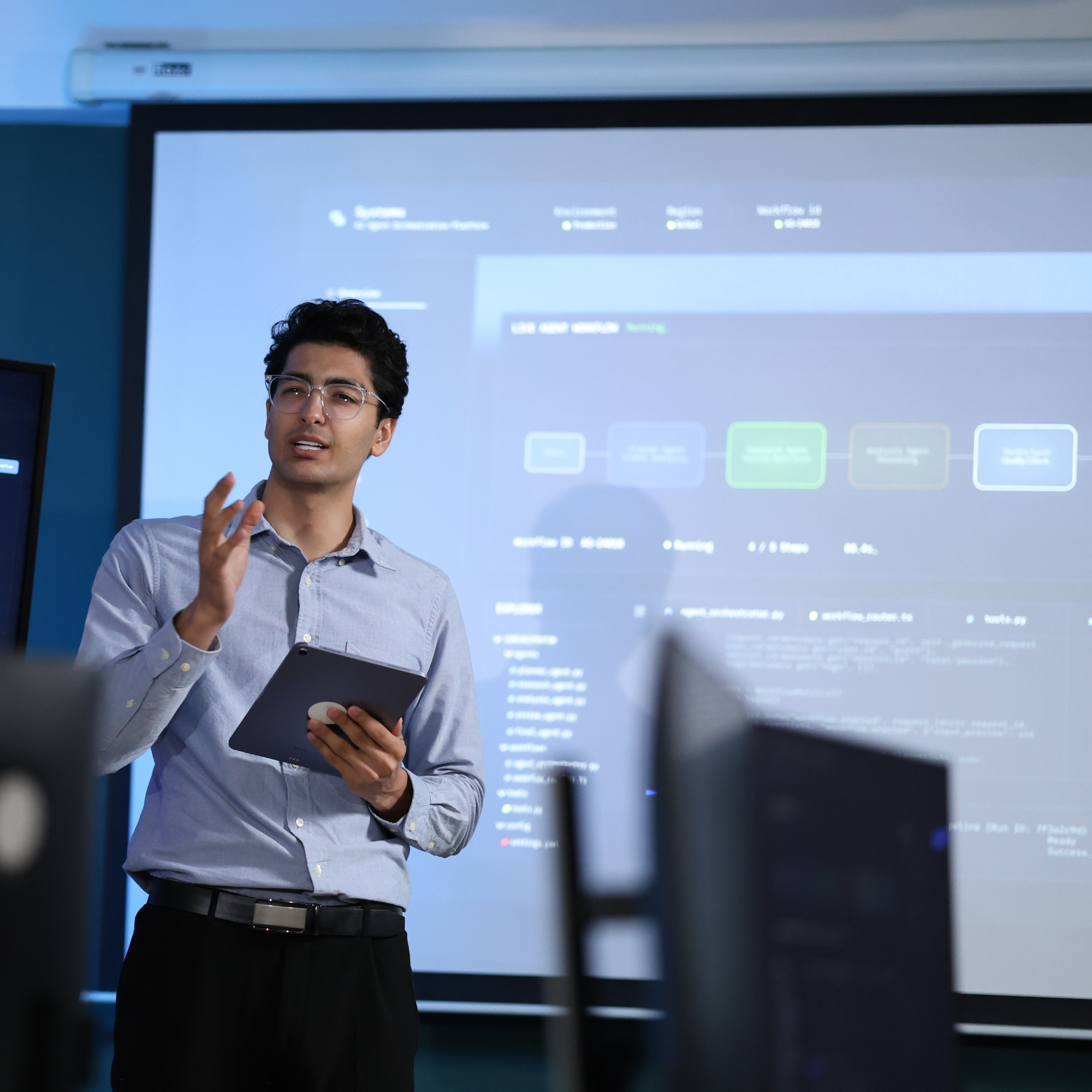 A presenter explaining a technical topic beside a projection screen.